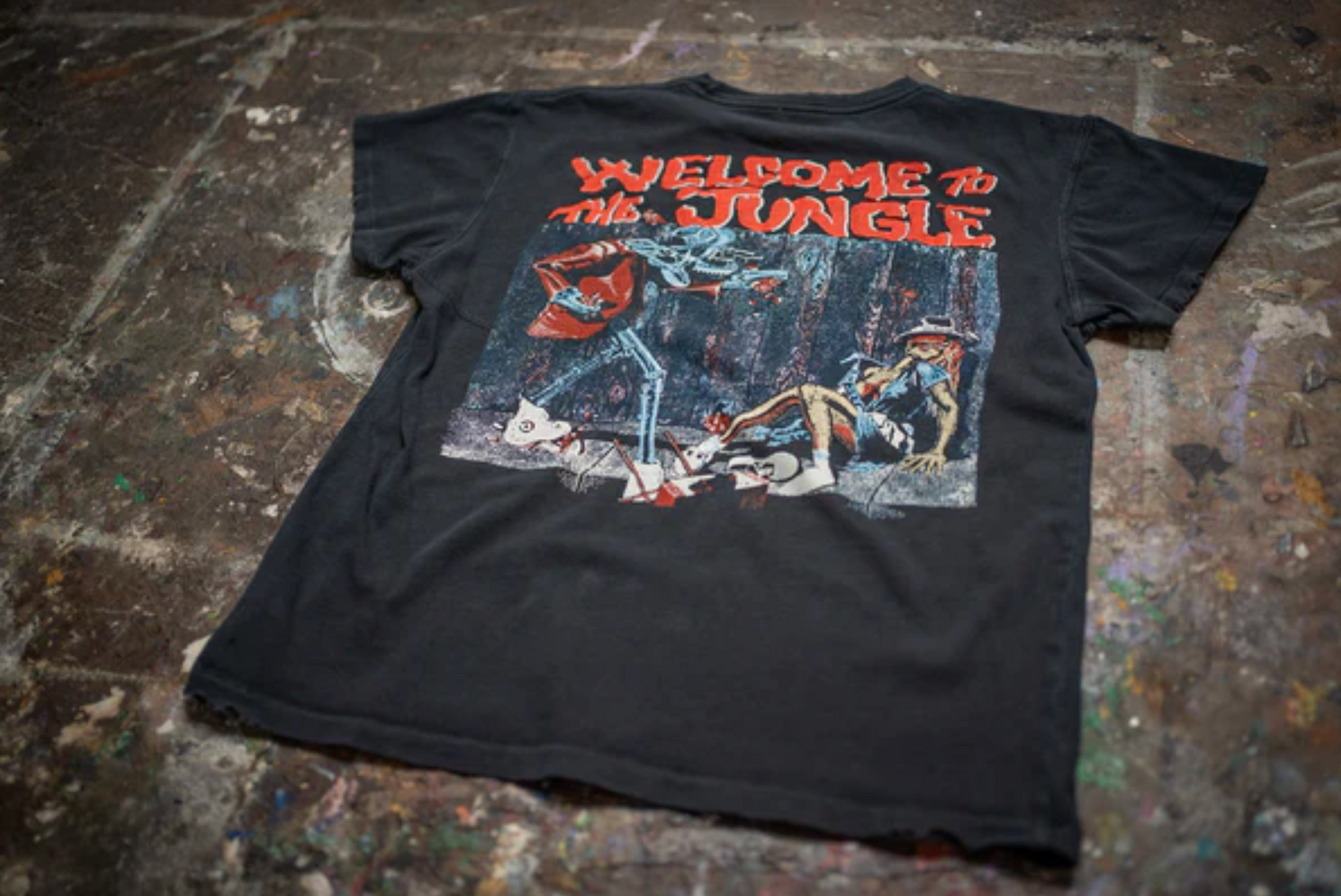 A black Guns N Roses Crew Tee by Made Worn rests on a paint-splattered surface. The back shows vivid skeleton and jungle artwork with bold red "WELCOME TO THE JUNGLE" text, inspired by Guns N Roses.
