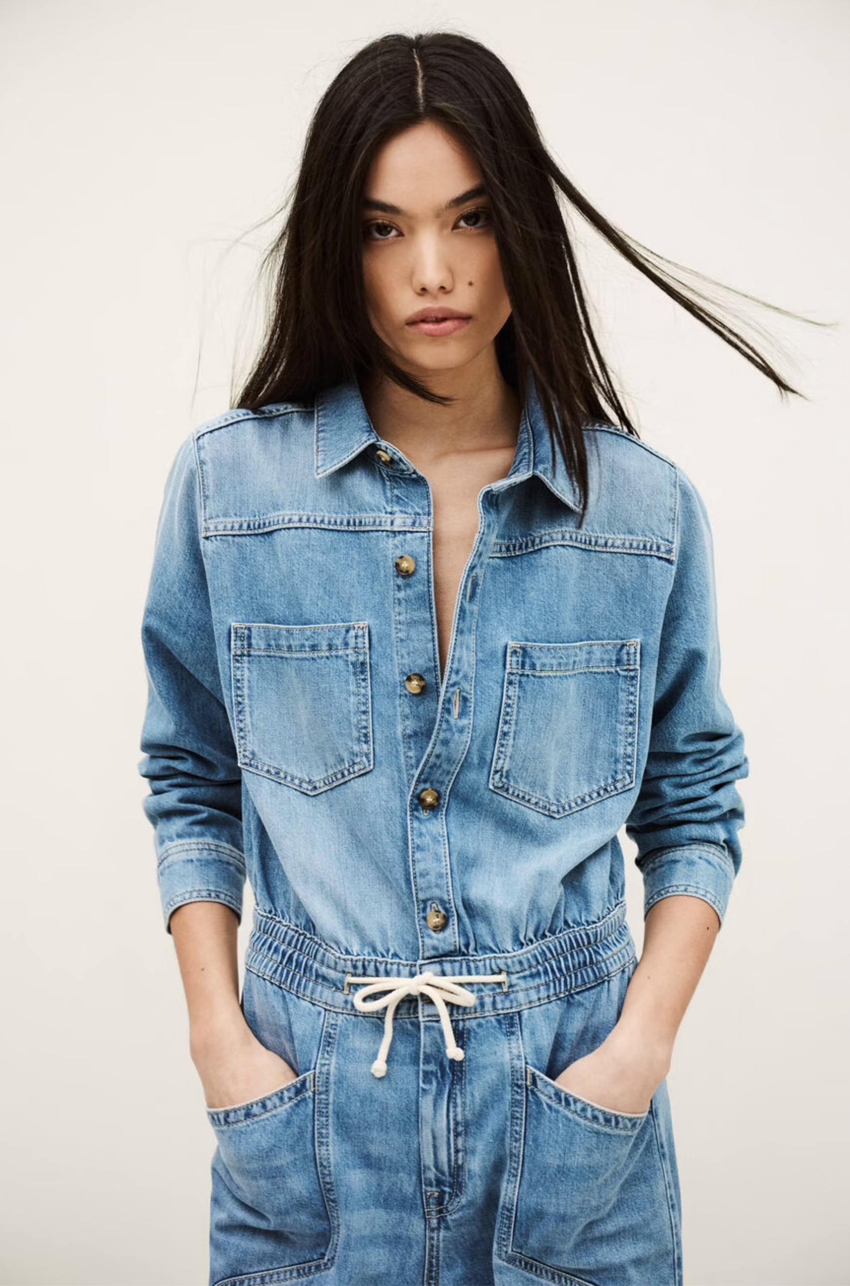 A woman with long dark hair wears the BA&SH Jumpsuit Mirella, a denim cargo style with utility pockets and a drawstring waist, standing against a plain light background with her hands in her pockets.