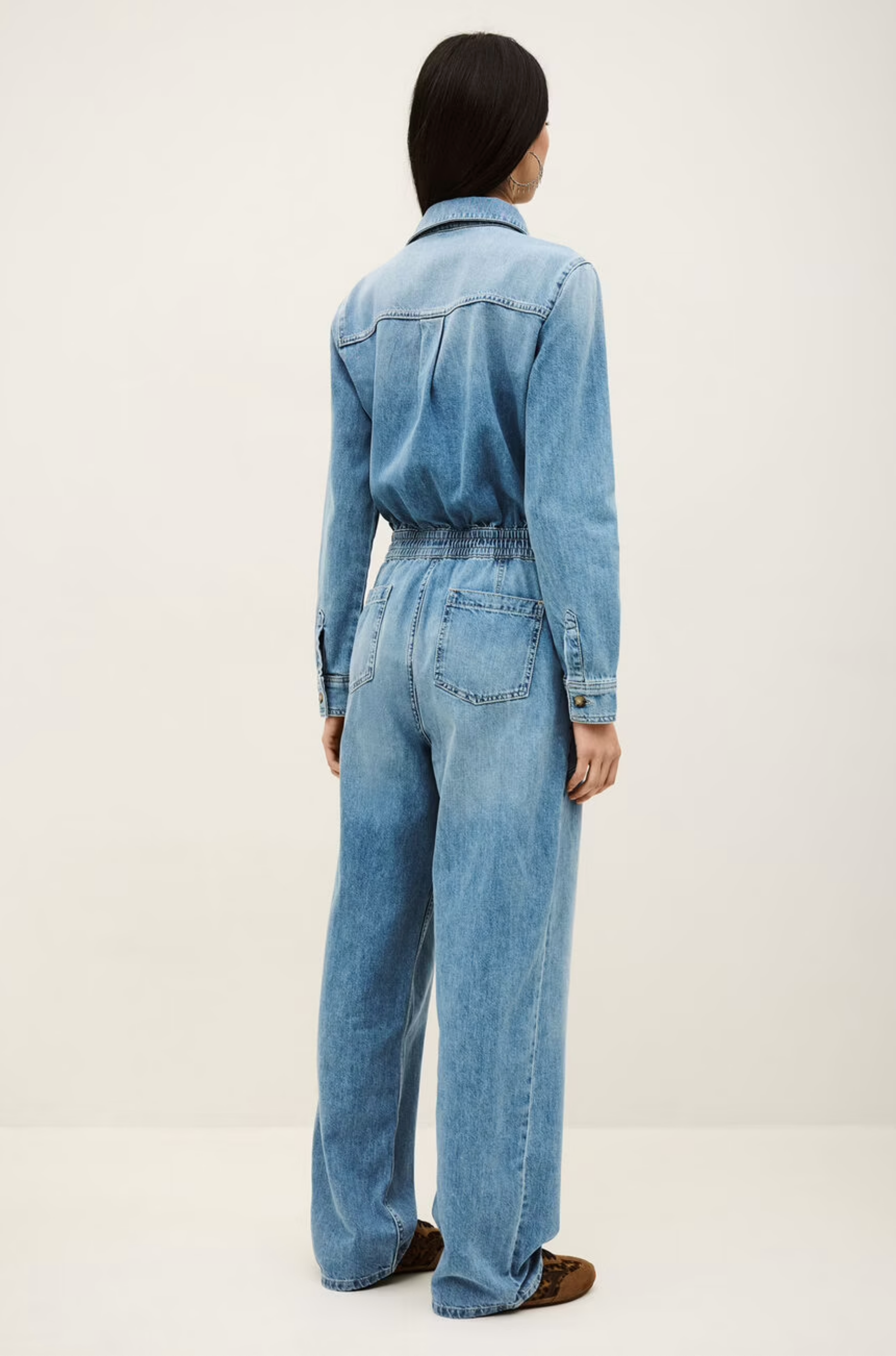 A person with long dark hair, seen from behind, wearing the BA&SH Jumpsuit Mirella—a light blue denim cargo style with utility pockets and wide legs—stands against a plain light background.