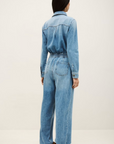 A person with long dark hair, seen from behind, wearing the BA&SH Jumpsuit Mirella—a light blue denim cargo style with utility pockets and wide legs—stands against a plain light background.