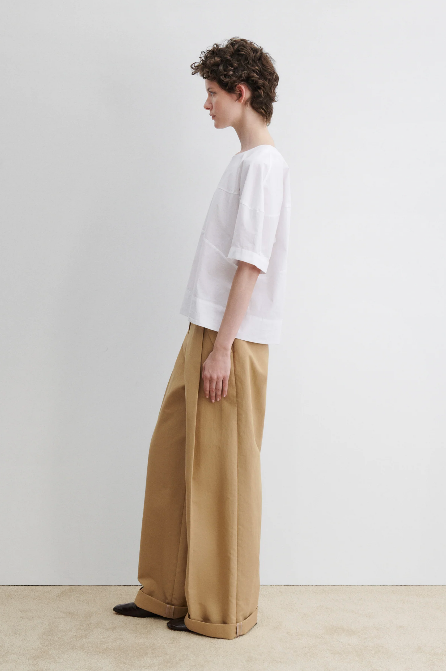 A person with short curly hair stands in profile against a white wall, wearing the Rachel Comey Rugby Top in organic Portuguese cotton and wide-legged tan pants with cuffs, paired with dark shoes.