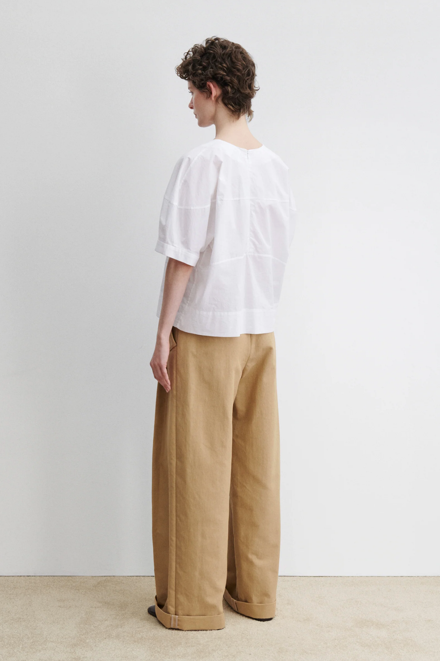 A person with short curly hair stands sideways, wearing the Rachel Comey Rugby Top and wide-legged, cuffed tan pants against a plain white wall and beige floor.