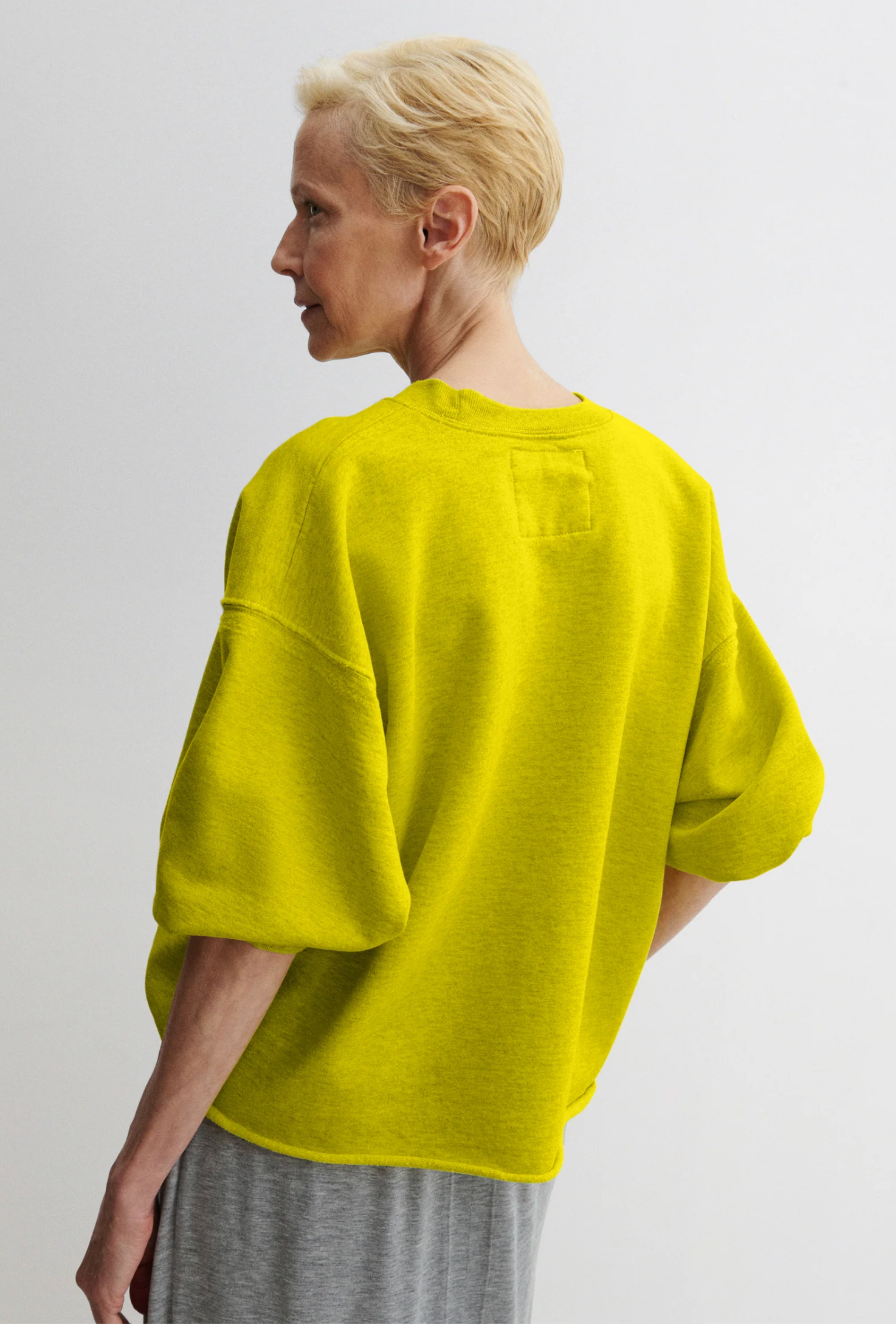 A person with short blonde hair, seen from behind, wears the Rachel Comey Fond Sweatshirt—a loose, bright yellow crewneck with wide cropped sleeves—paired with light gray pants against a plain light background.