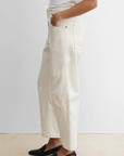 A person stands with one hand in their pocket against a plain background, wearing a white short-sleeve top, black slip-on shoes, and Rachel Comey’s cream-colored Elkin Pant featuring an adjustable back buckle.