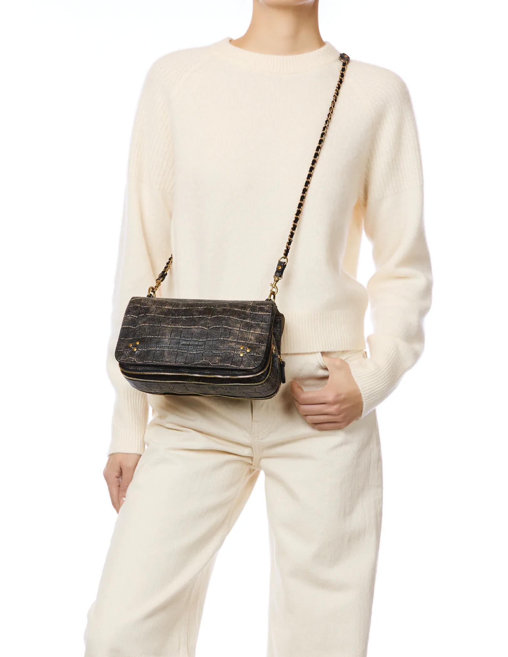 A person in a cream sweater and matching pants poses with the Bobi Bag by Jerome Dreyfuss, a black textured goatskin bag with an adjustable chain strap, against a plain white background.