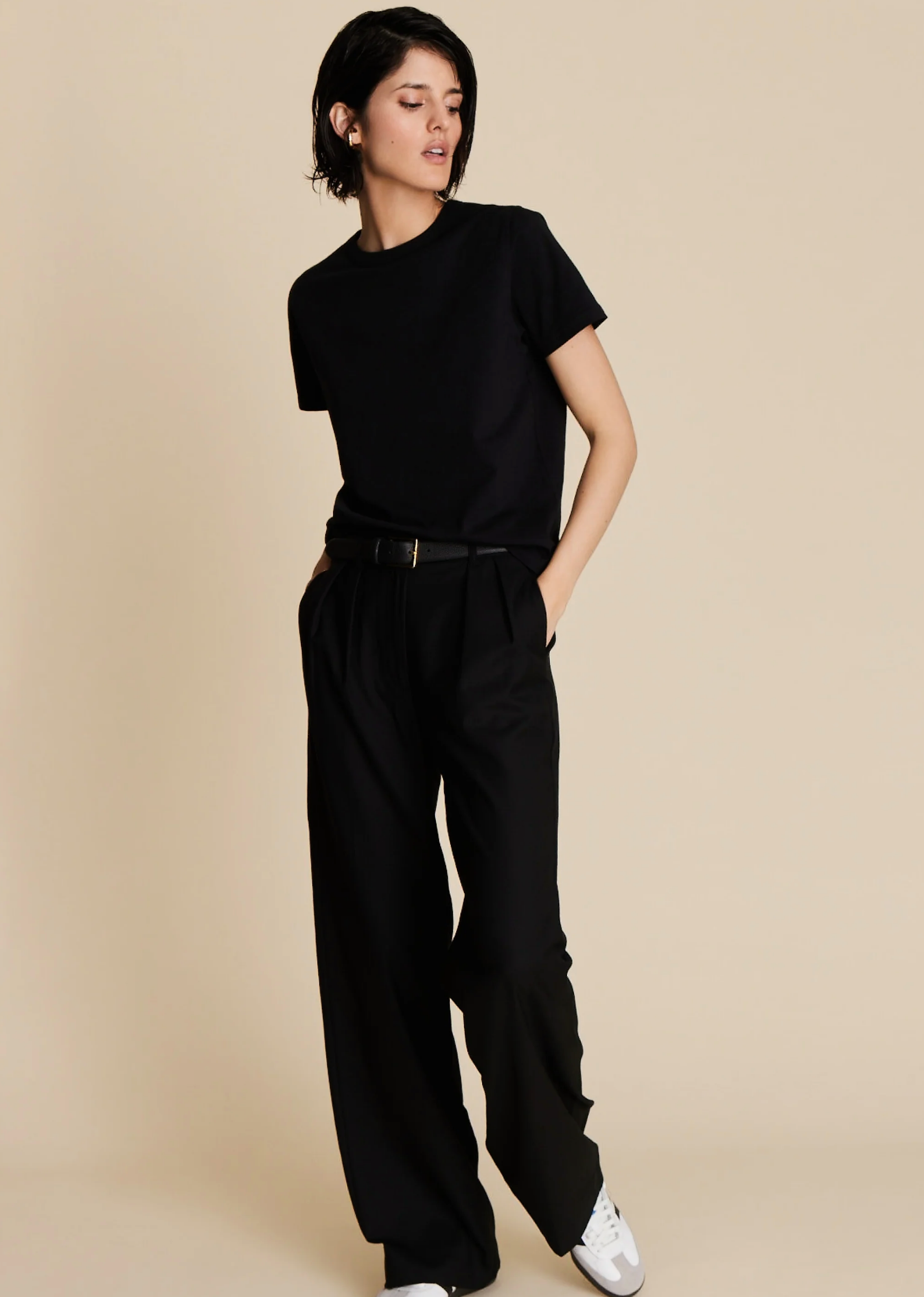 A person with short dark hair wears Sold Out NYC’s The Soft Perfect Tee in black, paired with belted black wide-leg trousers and white sneakers, standing against a plain beige background with one hand in their pocket.