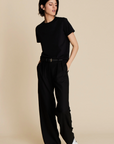 A person with short dark hair wears Sold Out NYC’s The Soft Perfect Tee in black, paired with belted black wide-leg trousers and white sneakers, standing against a plain beige background with one hand in their pocket.
