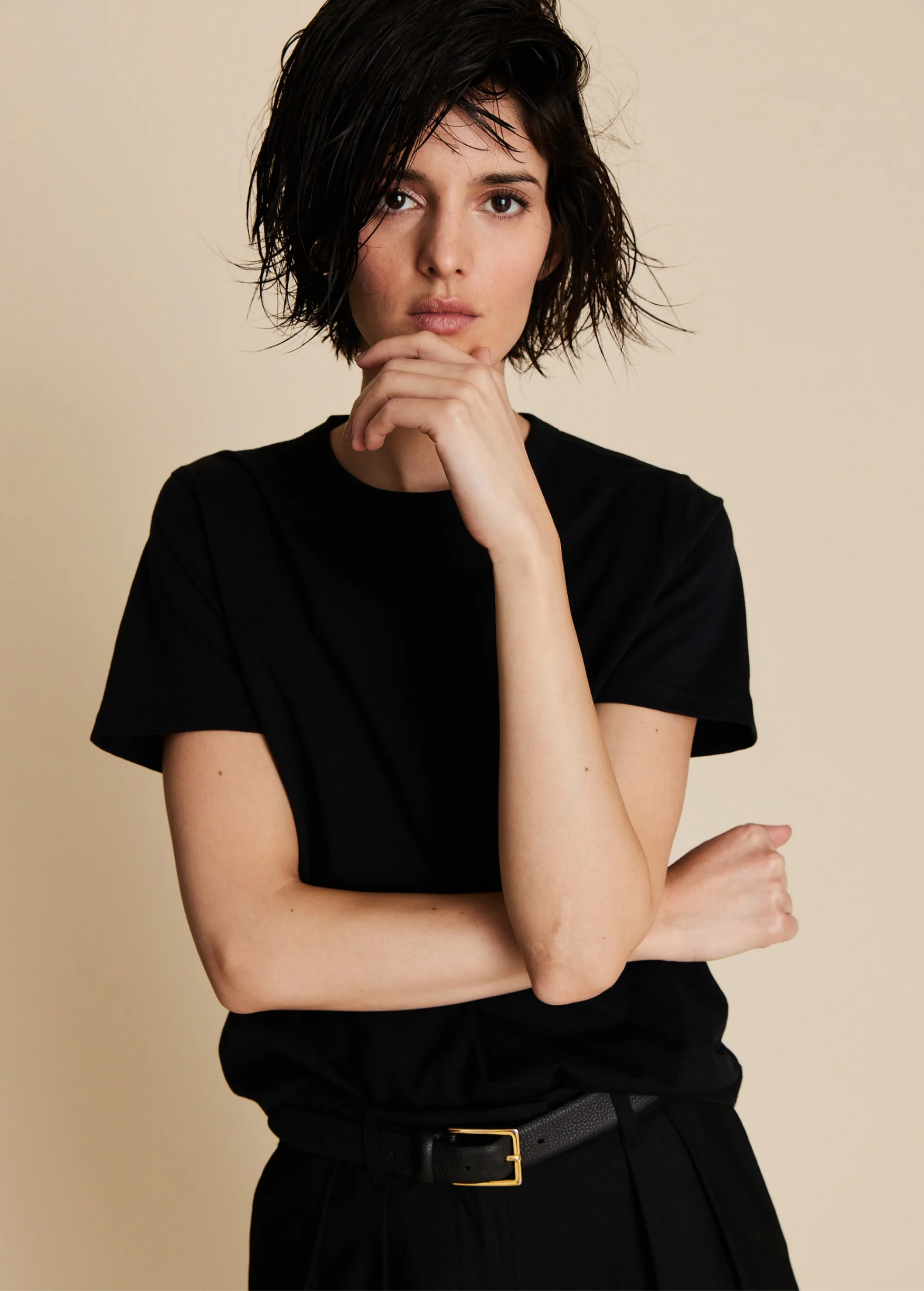 A person with short, tousled dark hair models The Soft Perfect Tee by Sold Out NYC—a classic black crew neck—paired with a belt against a beige background, one hand on their chin and the other arm crossed.