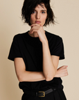 A person with short, tousled dark hair models The Soft Perfect Tee by Sold Out NYC—a classic black crew neck—paired with a belt against a beige background, one hand on their chin and the other arm crossed.