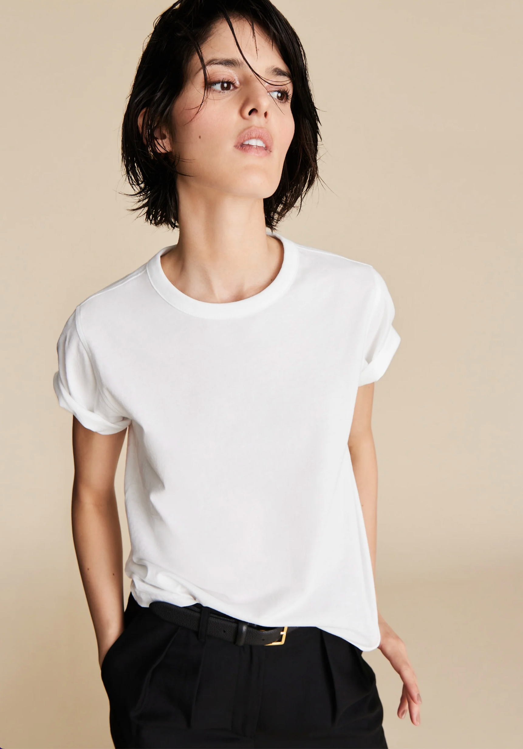 A person with short dark hair wears The Soft Perfect Tee from Sold Out NYC in organic Peruvian cotton and black pants, standing against a beige background and looking to the side with a relaxed expression.