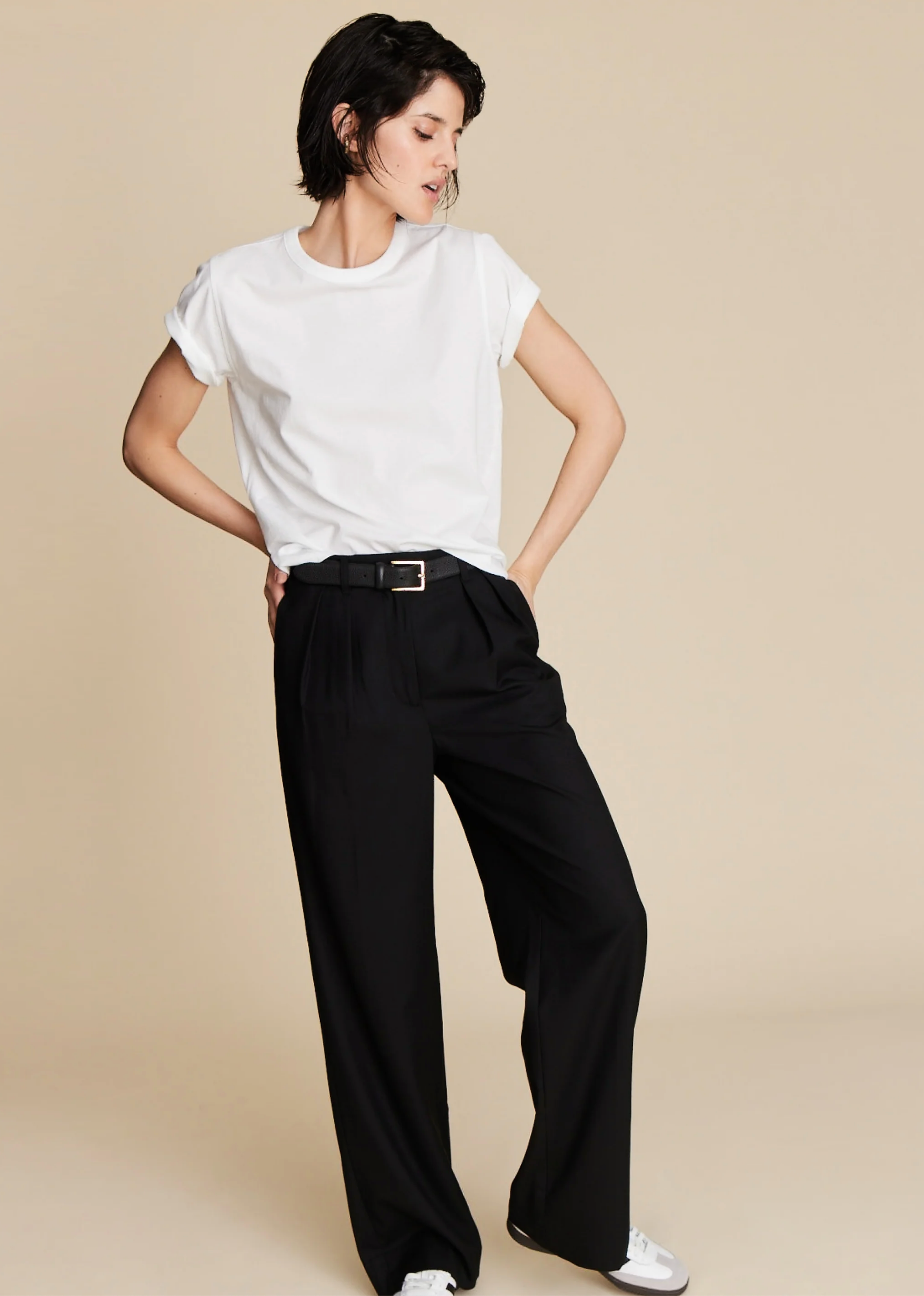 A person with short dark hair poses against a beige background, wearing Sold Out NYC’s The Soft Perfect Tee—an organic Peruvian cotton crew neck—and loose black high-waisted belted trousers with white sneakers.