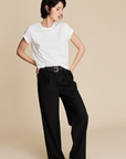 A person with short dark hair poses against a beige background, wearing Sold Out NYC’s The Soft Perfect Tee—an organic Peruvian cotton crew neck—and loose black high-waisted belted trousers with white sneakers.