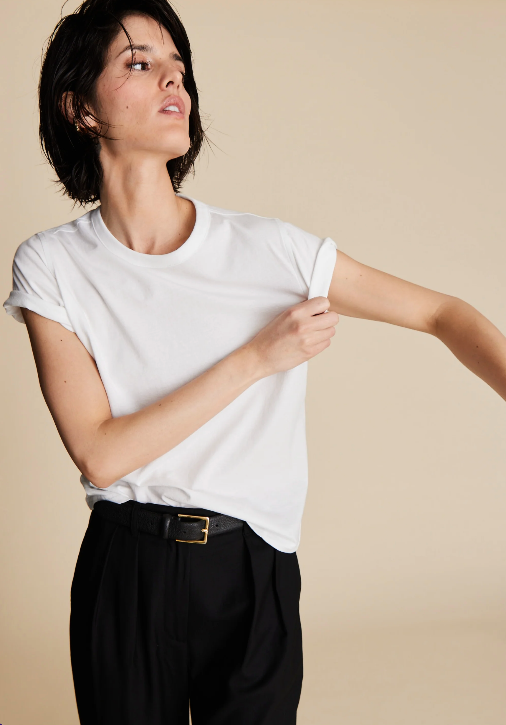 A person with short dark hair wears The Soft Perfect Tee by Sold Out NYC, a relaxed fit shirt made from organic Peruvian cotton, tucked into black belted trousers while adjusting their shirt against a neutral beige backdrop.