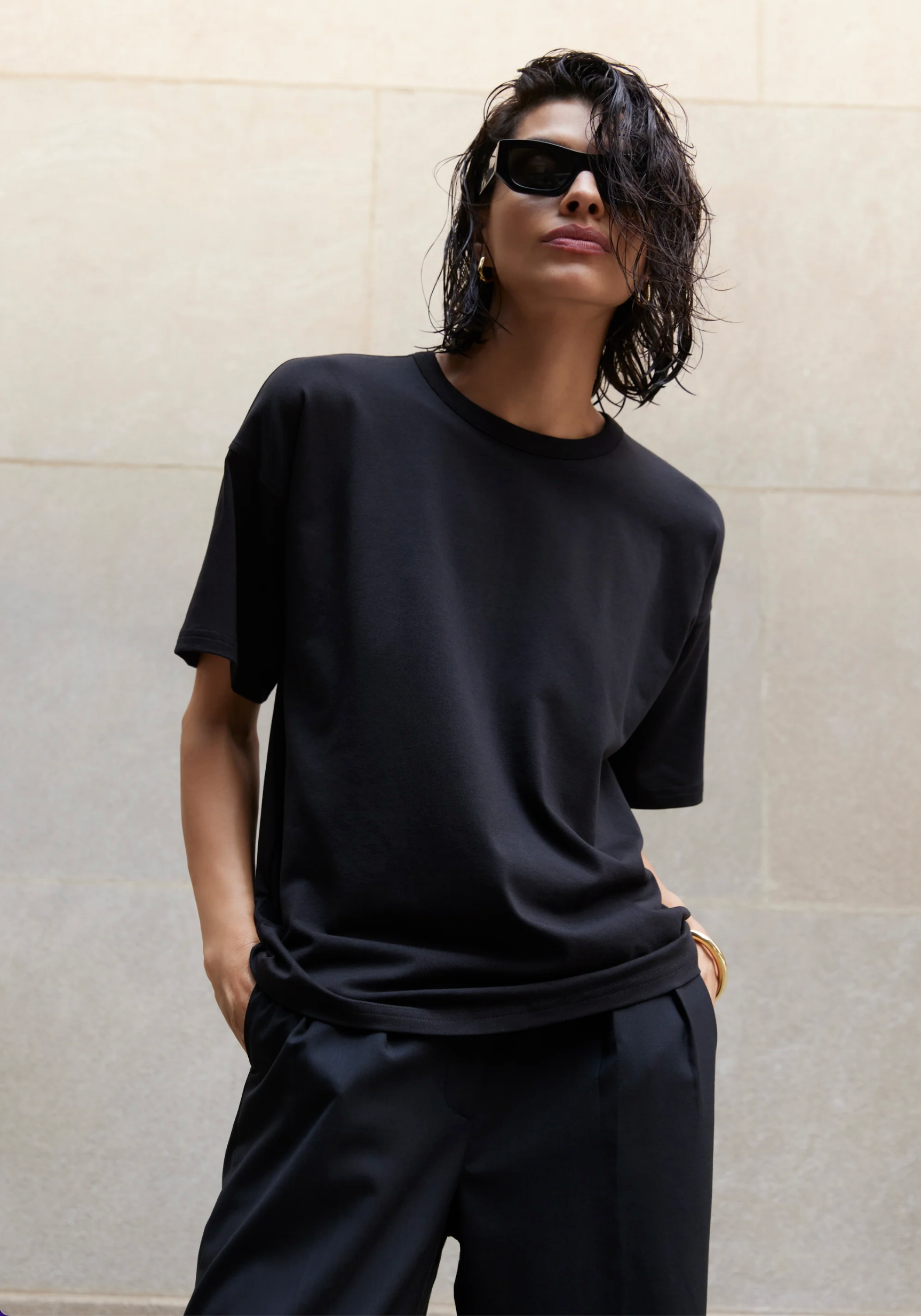 A person with wet, chin-length dark hair wears black sunglasses, black pants, and Sold Out NYC's The Oversized Tee—known for its buttery-soft feel—standing by a light stone wall with one hand in their pocket.