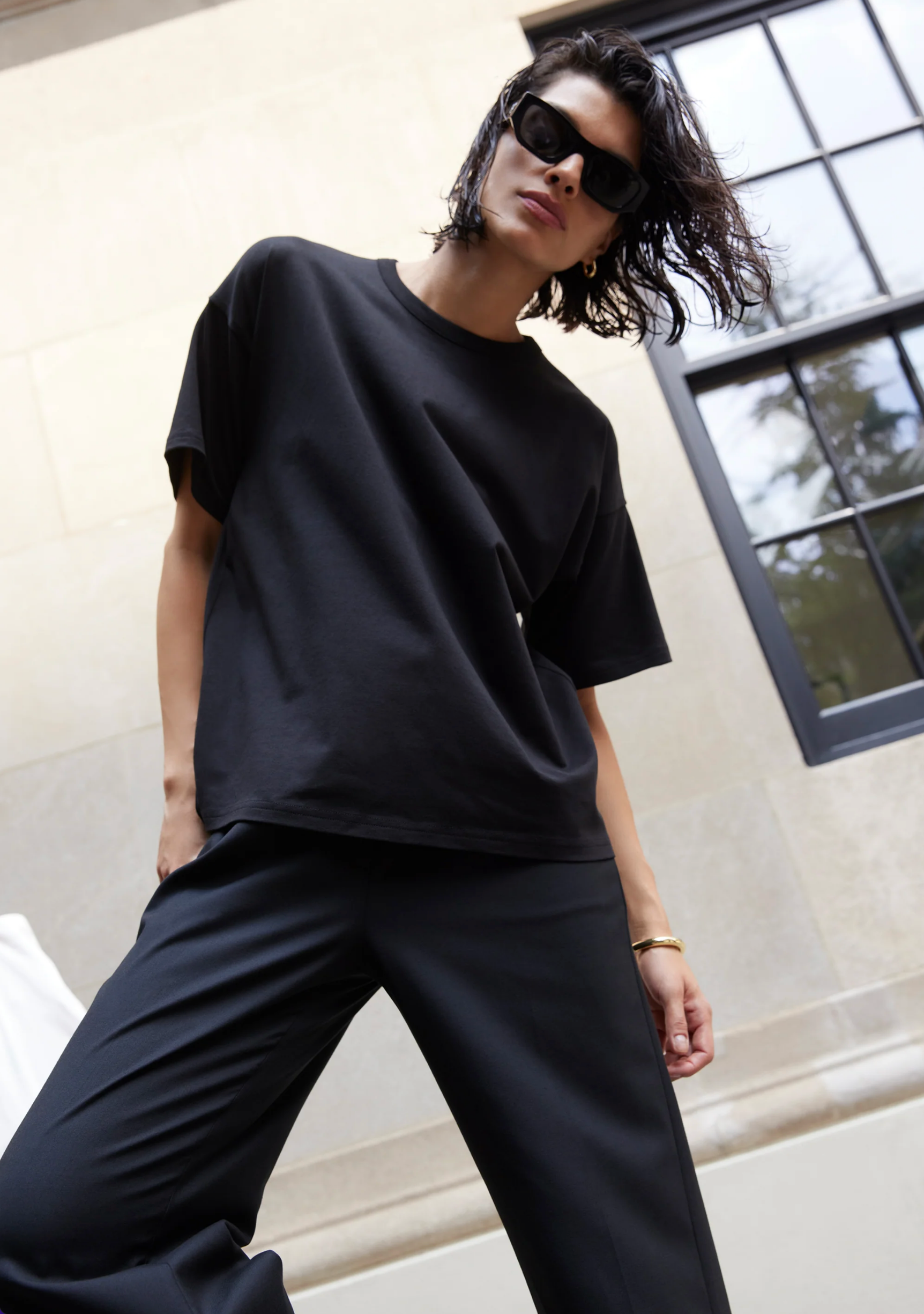 A person with short, wavy hair wearing black sunglasses, black pants, and The Oversized Tee by Sold Out NYC stands outdoors in front of a light stone building with a large window, leaning slightly forward.