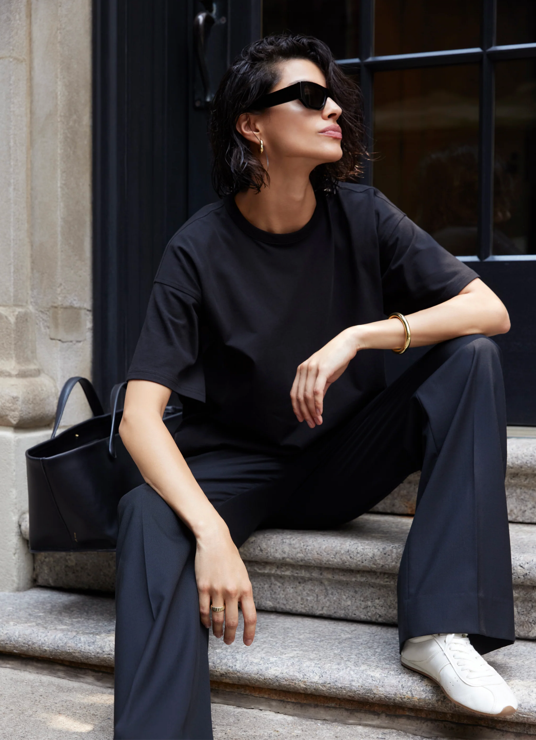 A person with short dark hair and sunglasses sits on stone steps, wearing Sold Out NYC’s The Oversized Tee in black, paired with black pants and white sneakers. A black handbag rests beside them.