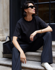 A person with short dark hair and sunglasses sits on stone steps, wearing Sold Out NYC’s The Oversized Tee in black, paired with black pants and white sneakers. A black handbag rests beside them.