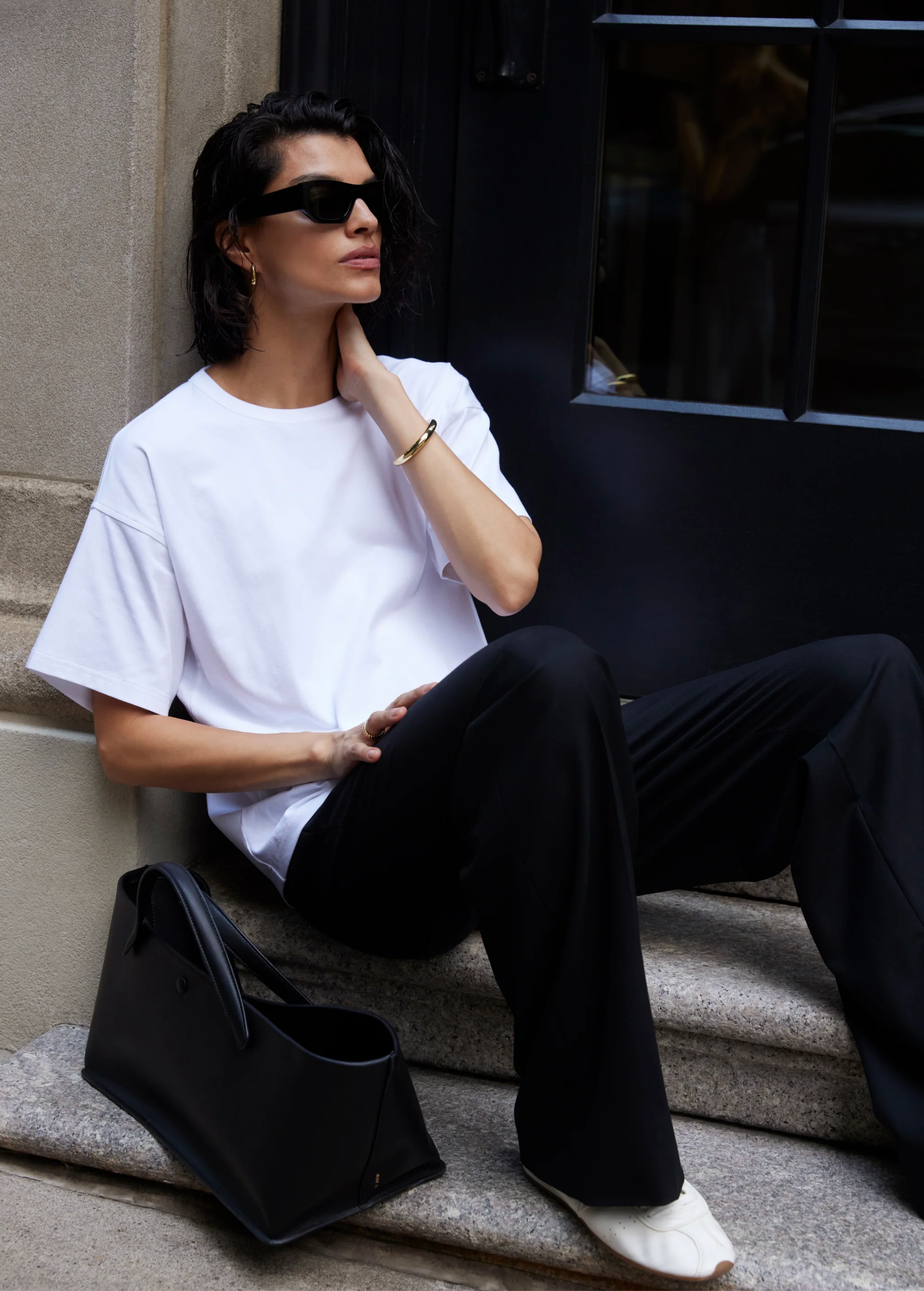 A person with shoulder-length dark hair and sunglasses sits on stone steps beside a black bag, wearing Sold Out NYC’s The Oversized Tee—crafted from buttery-soft heavyweight cotton—and black pants, resting a hand on their neck and looking aside.