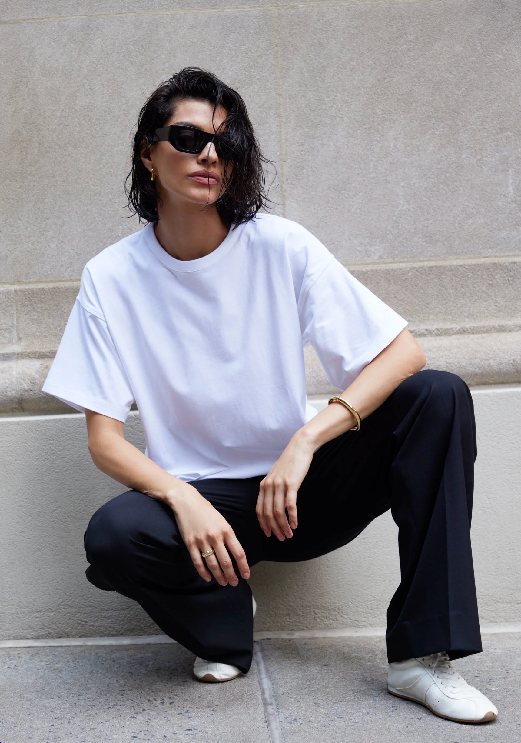 A woman with short dark hair and black sunglasses wears Sold Out NYC’s The Oversized Tee, black pants, and white sneakers as she squats confidently against a light stone wall.