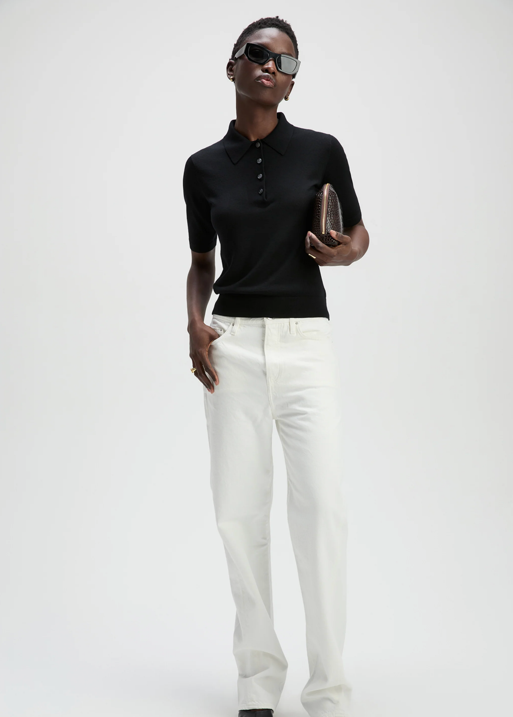 Wearing sunglasses, The Refined SS Polo by Sold Out NYC as a women's layering piece, and white pants, a person stands holding a textured clutch against a plain light background.
