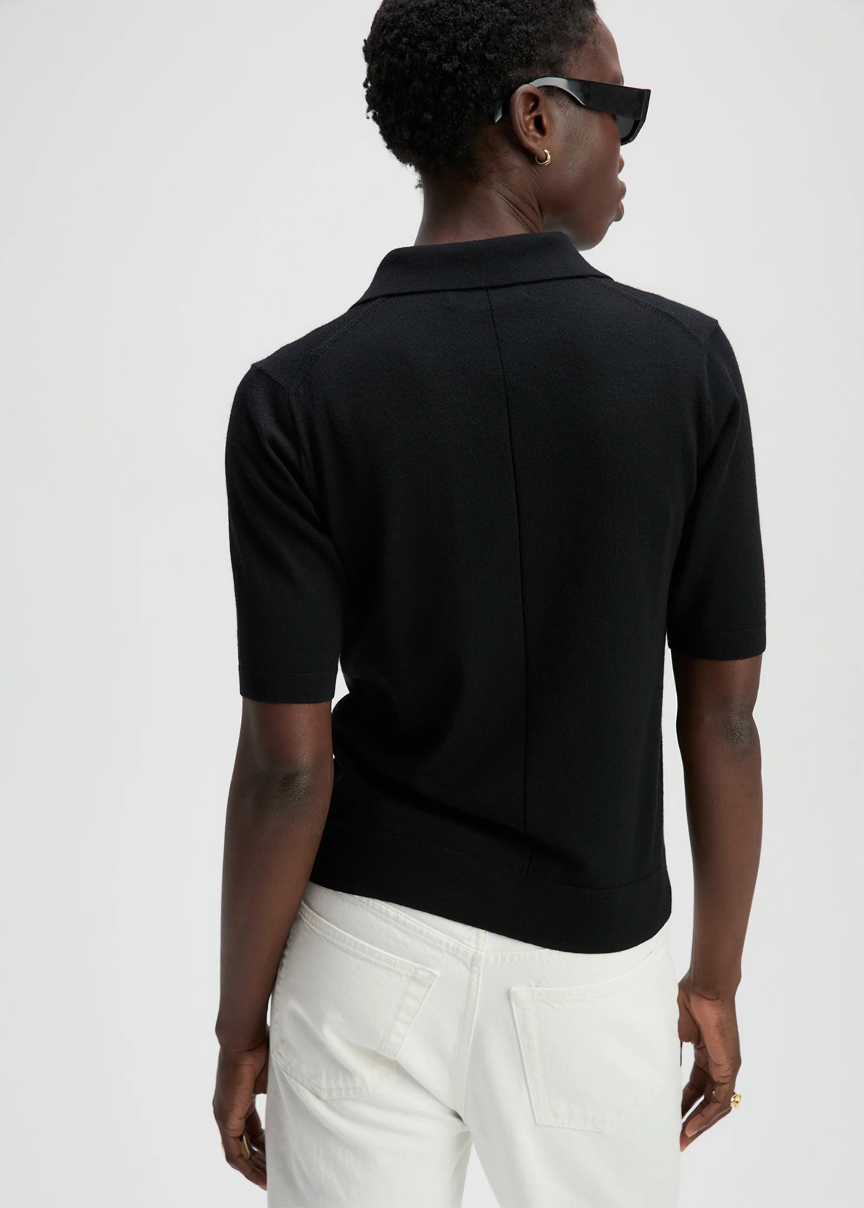 Someone wearing The Refined SS Polo by Sold Out NYC and white pants is seen from behind, with short hair and dark sunglasses, standing against a plain light background.