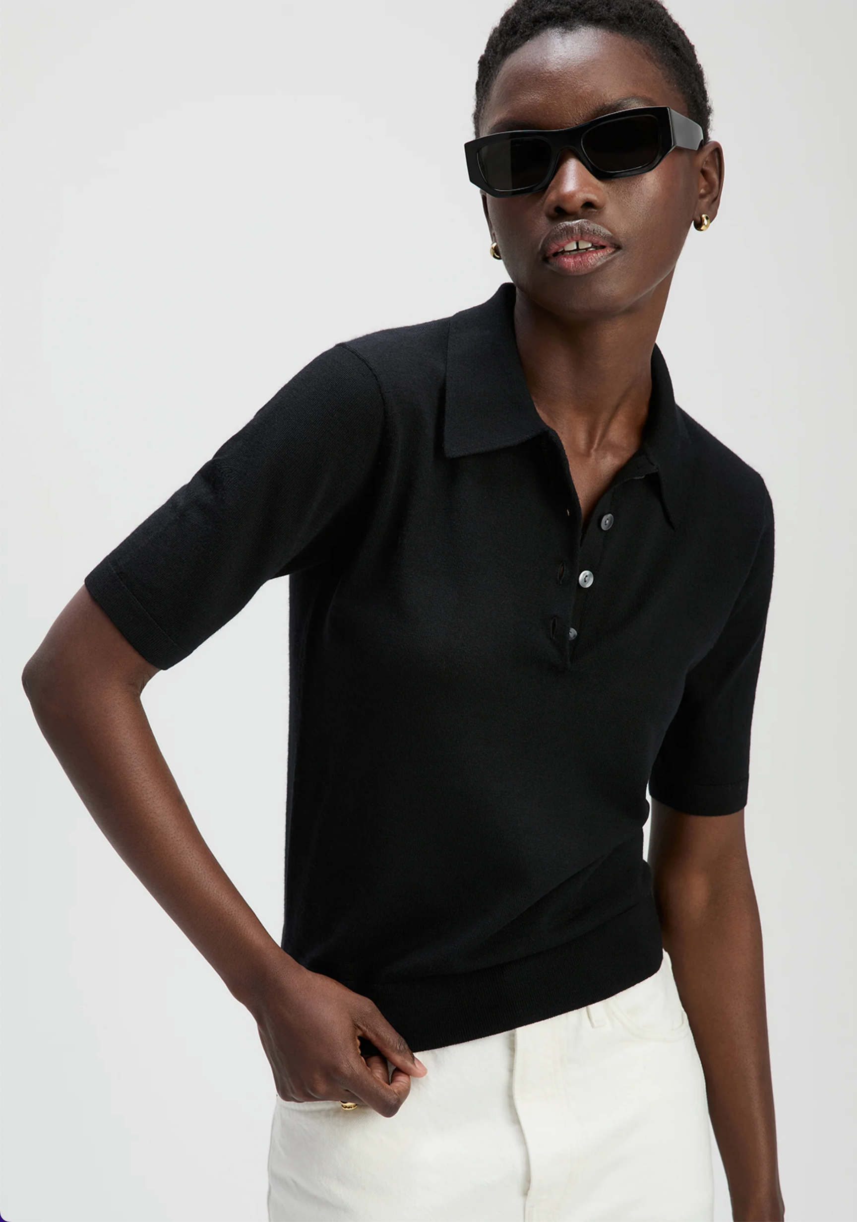 A person wears Sold Out NYC’s The Refined SS Polo—a black, short-sleeve collared shirt with buttons—paired with white pants and black sunglasses, posing against a plain light background.