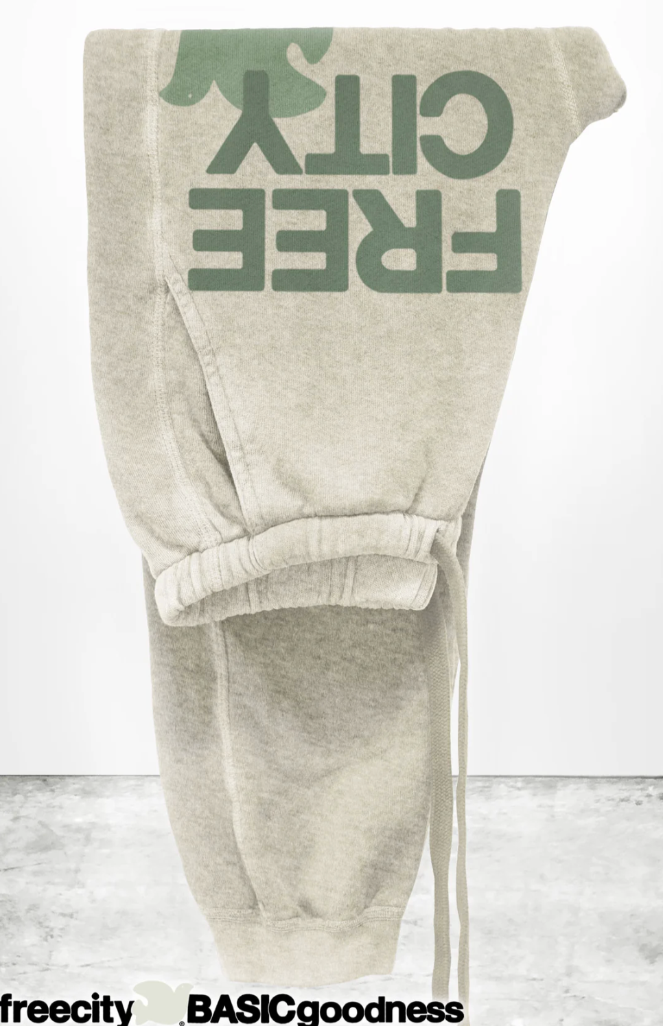 The FREECITY LARGE SUNFADES pocket sweatpant by Free City (sparrow, LLC) is a light beige unisex French terry pant shown upside down, with "FREE CITY" in large green letters and "freecity BASICgoodness" in black on the leg.