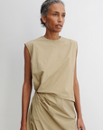 A woman with curly gray hair pulled back is wearing the Rachel Comey Rini Dress, an asymmetrical sleeveless beige dress with a matching draped skirt, standing against a plain white background.
