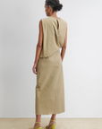 A woman with curly hair in a low bun stands with her back to the camera, wearing the Rachel Comey Rini Dress and yellow heeled sandals against a plain white background.