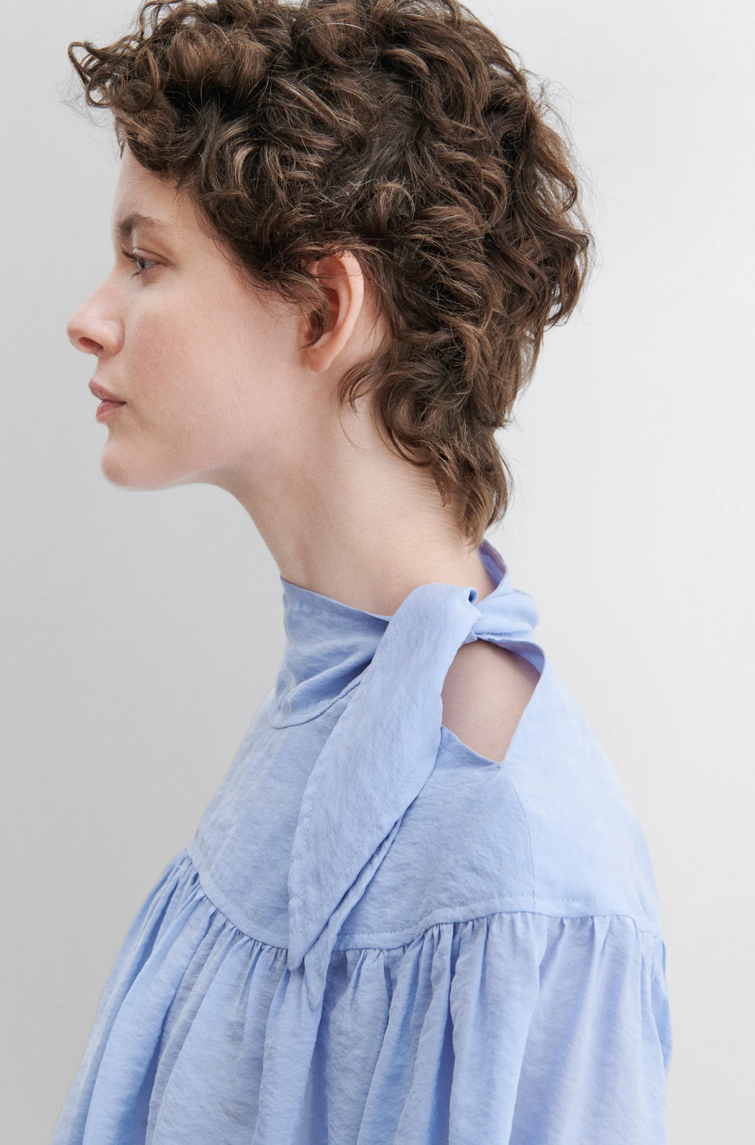 A person with short, curly brown hair is shown in profile wearing the Rachel Comey Parachute Top—a light blue, textured, wrinkle-resistant long-sleeve blouse featuring a high collar and bow detail—against a plain, light background.