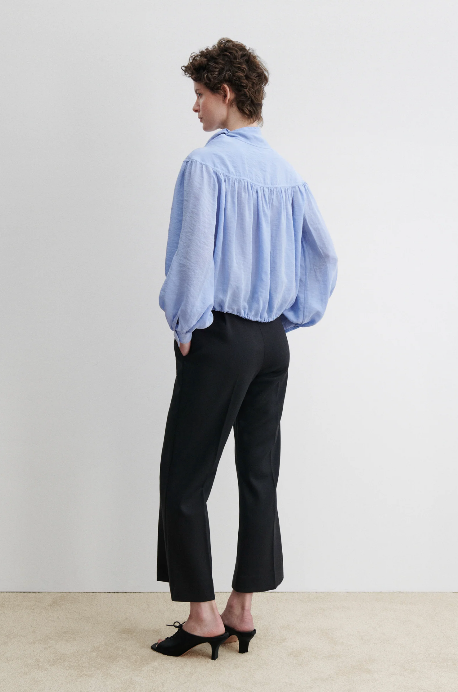 A person stands with hands in pockets, wearing the Rachel Comey Parachute Top— a loose light blue long-sleeve blouse— paired with black wide-leg trousers and heeled shoes, set against a plain white background.