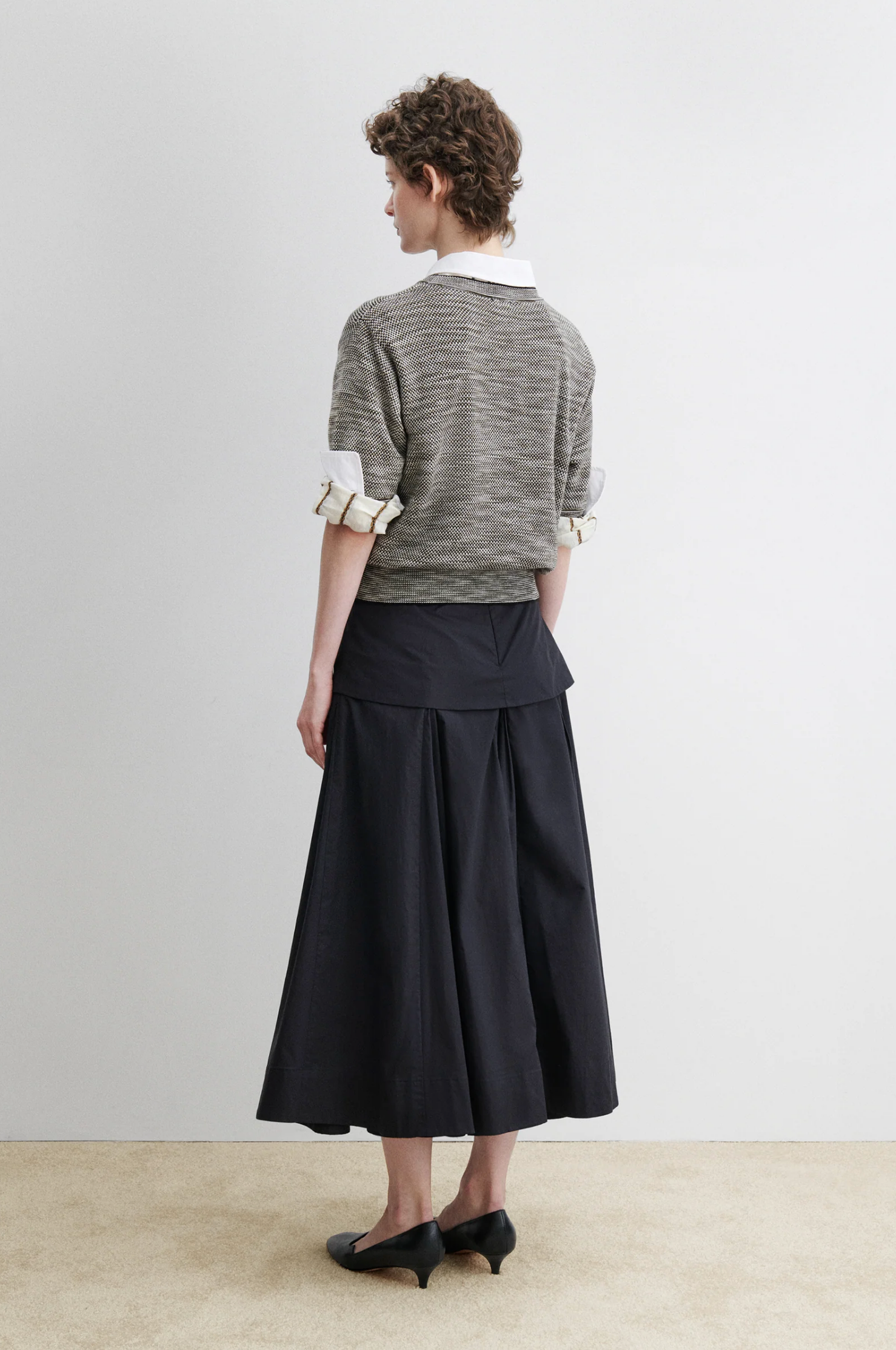 A person faces away wearing the Rachel Comey Jeram Top, a gray cotton tweed sweater with a crew neckline over a white collared shirt, paired with a long black skirt and black shoes against a plain light background.