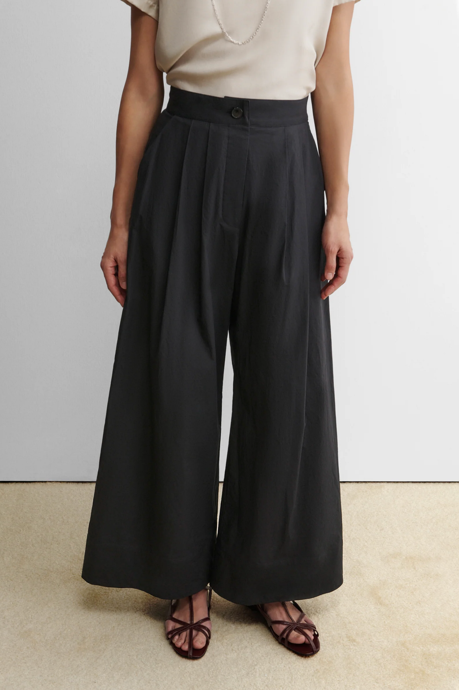 Someone stands against a plain background wearing Rachel Comey's Coxsone Pant—high-waisted, wide leg, black organic cotton—paired with a light short-sleeve top and brown open-toe sandals.