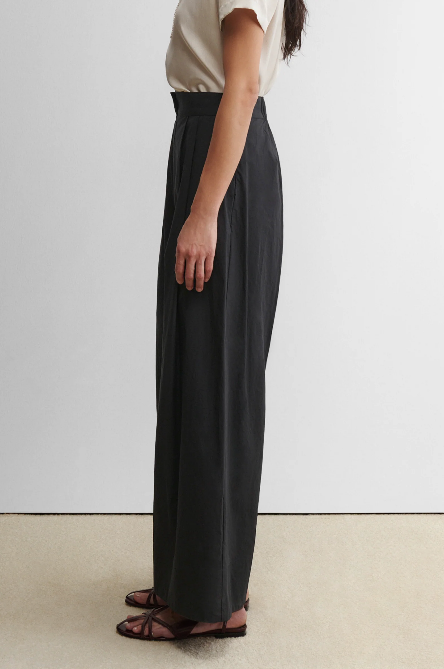 A person stands in profile against a plain background, wearing a short-sleeve beige top, brown flat sandals, and high-waisted wide leg Coxsone Pants by Rachel Comey in black.