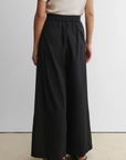 A person stands indoors facing away, wearing the Rachel Comey Coxsone Pant—high-waisted, wide-leg black trousers—paired with a short-sleeved beige top and sandals against a plain white wall background.