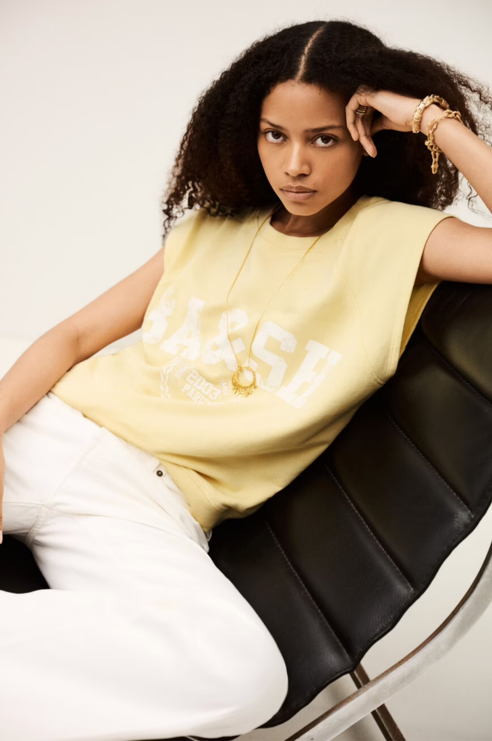 A woman with curly hair sits on a black leather chair, wearing the BA&SH Sweat Benta—a pale yellow sleeveless sweatshirt with American-style raglan armholes—paired with white pants and gold jewelry, looking directly at the camera with a serious expression.