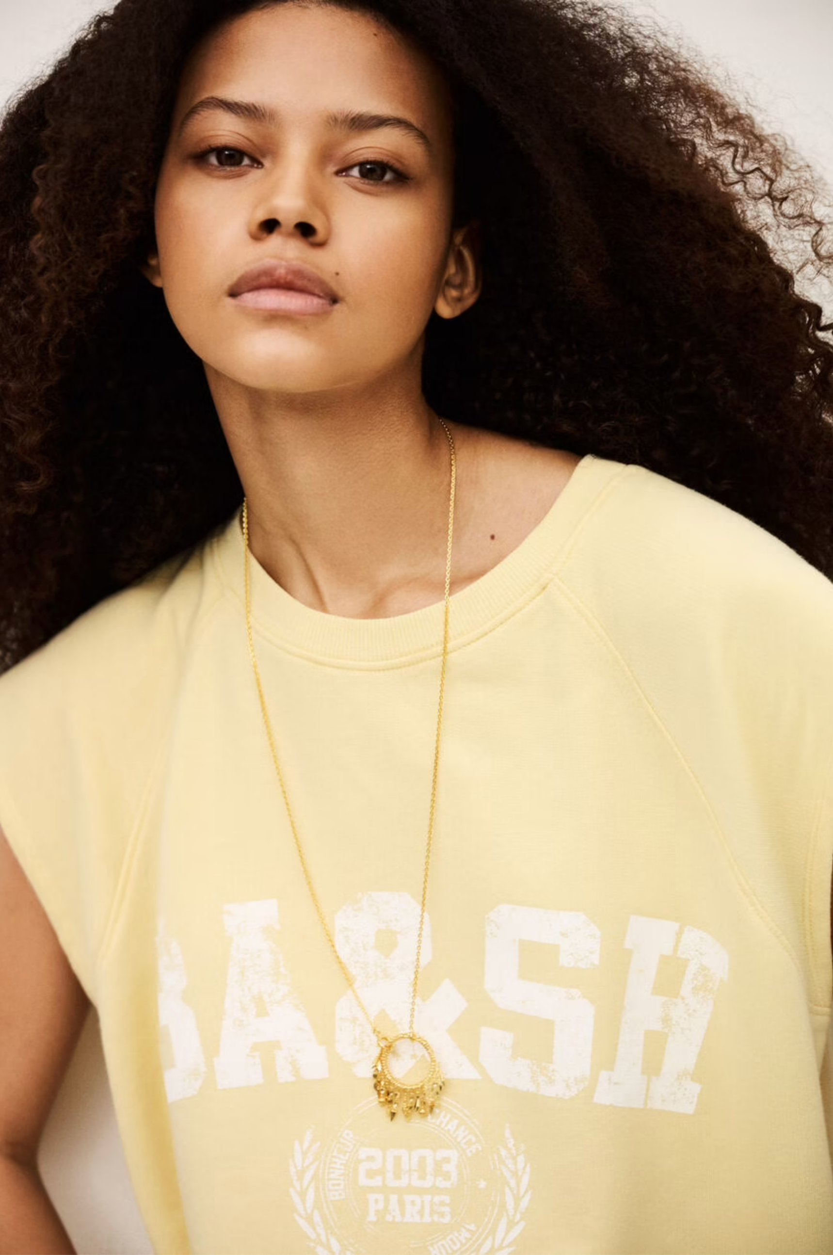 A young woman with voluminous curly hair wears the BA&SH Sweat Benta, a light yellow sleeveless sweatshirt with a round neckline and "BA&SH 2003 PARIS" print, accessorized with a long gold necklace, looking confidently at the camera.