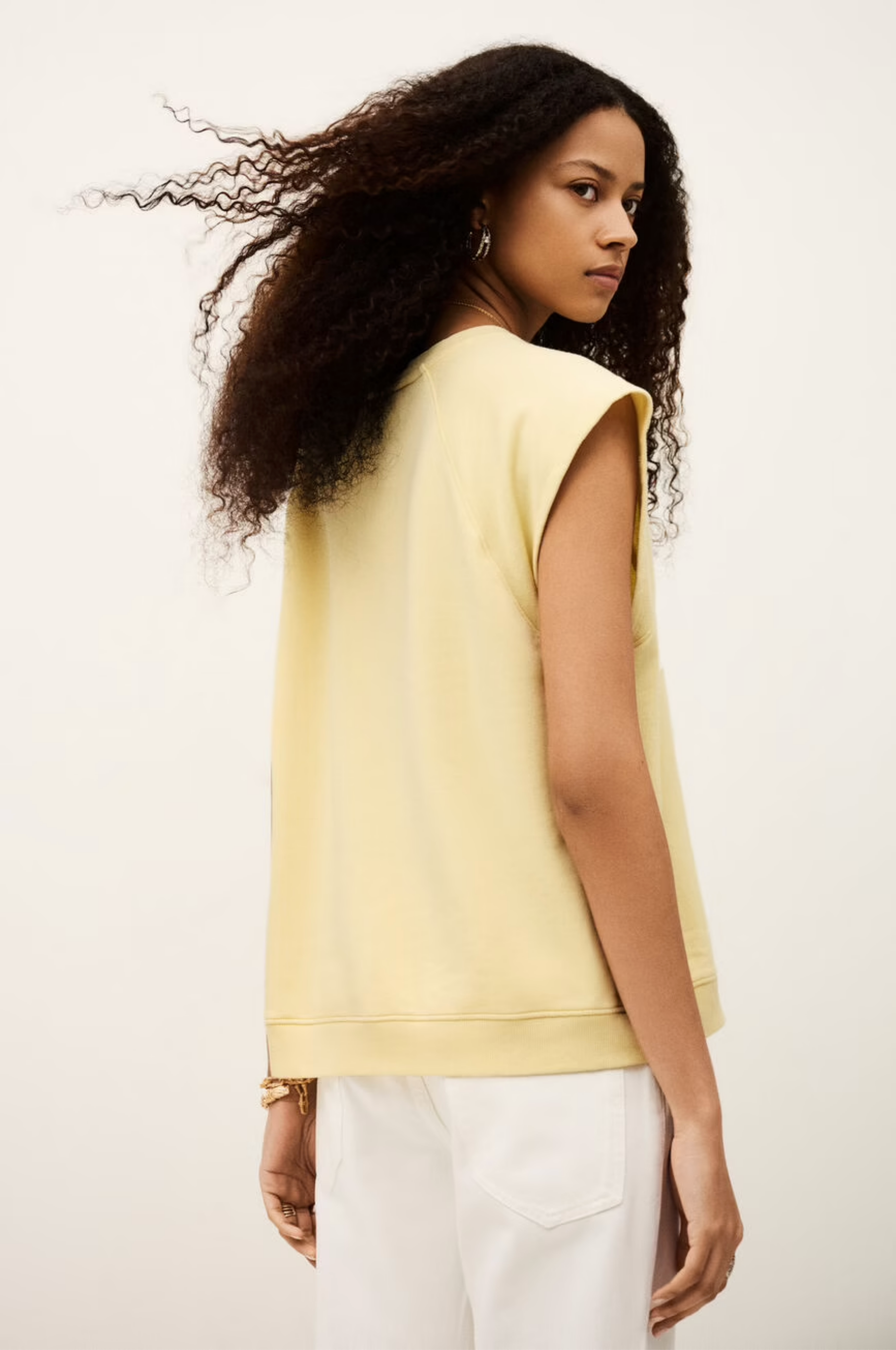 A woman with long curly hair models the BA&SH Sweat Benta, a sleeveless light yellow top with a round neckline, paired with white pants. She stands back to the camera, looking over her shoulder against a plain background.