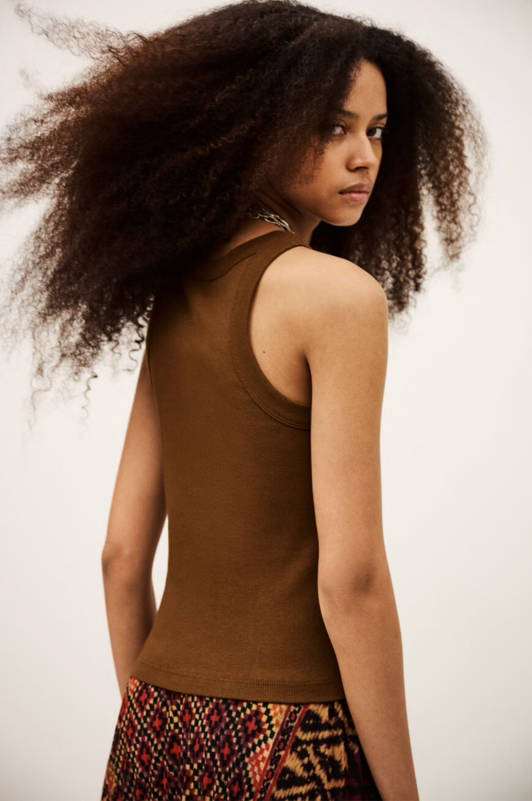 A young woman with voluminous curly hair captures modern boho style in the sleeveless brown BA&SH Top Cynda and a colorful patterned skirt, standing sideways and glancing over her shoulder against a plain backdrop.
