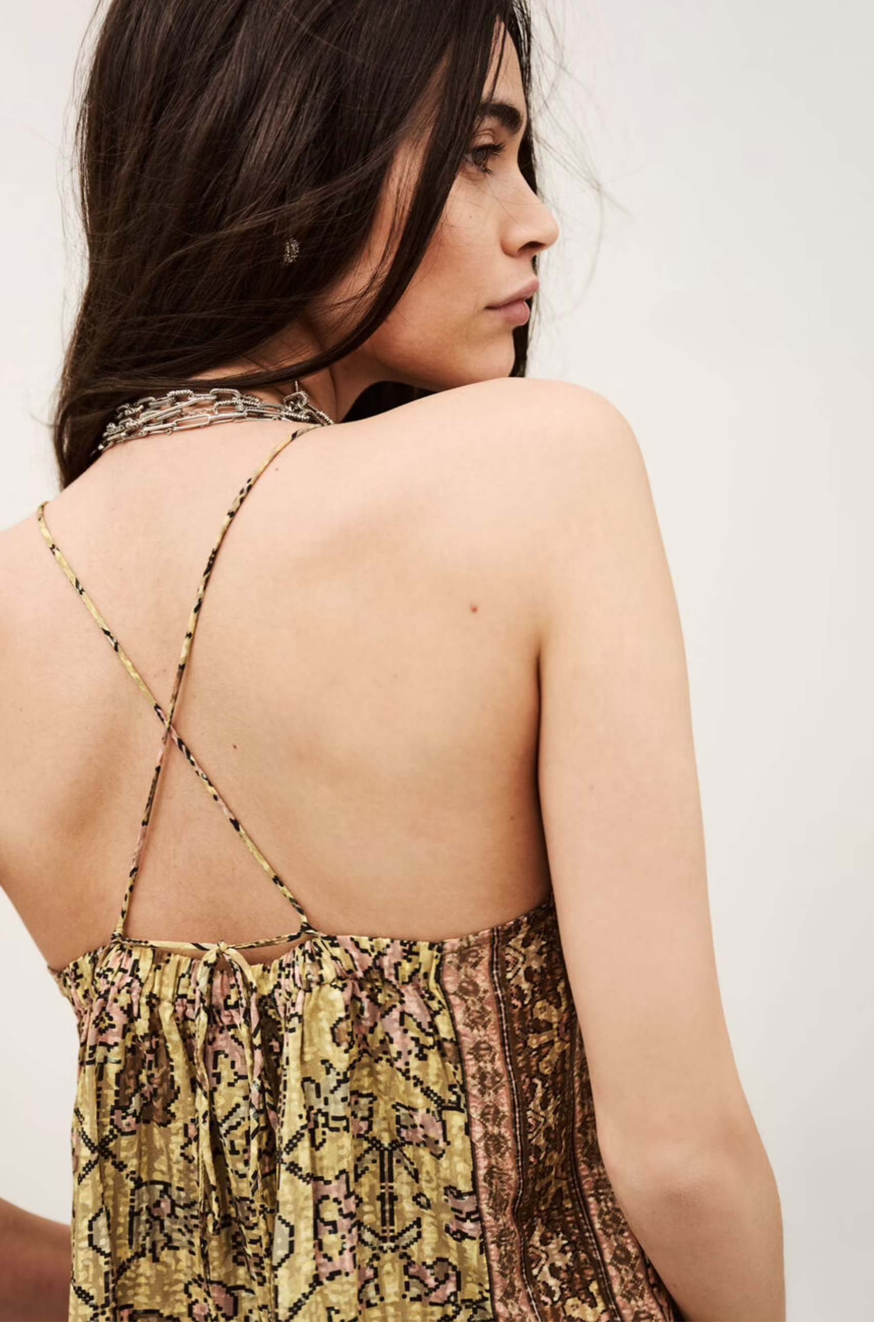 A woman with long dark hair is shown from the back, wearing the sleeveless Top Pamela by BA&SH. The top features thin, cross straps and an earthy-toned patterned fabric as she looks over her shoulder against a plain background.