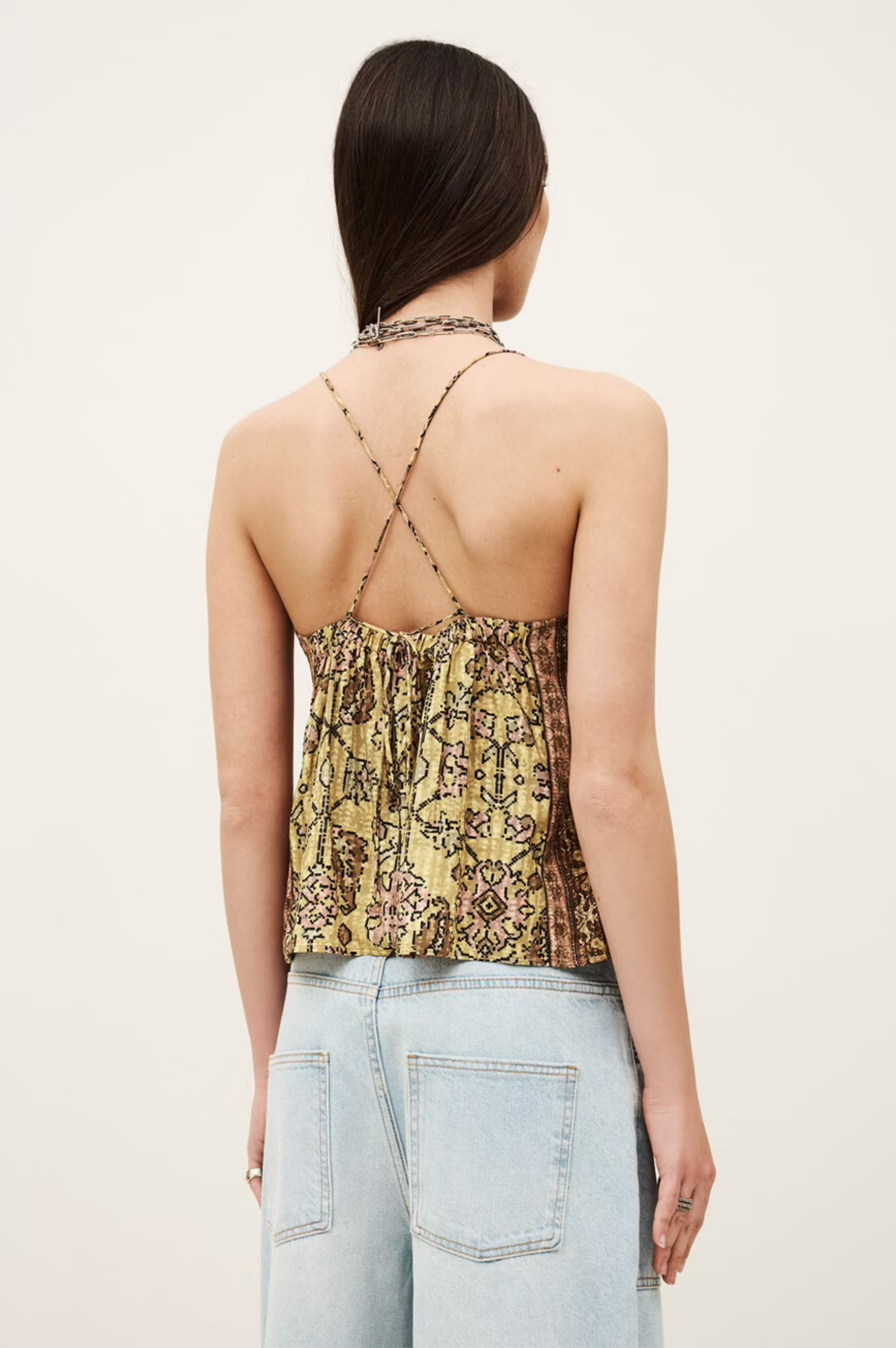 A woman with long dark hair models the BA&SH Top Pamela—a yellow and brown patterned halter crop top with metallic bead details and open back—paired with light blue high-waisted jeans, facing away from the camera against a plain background.