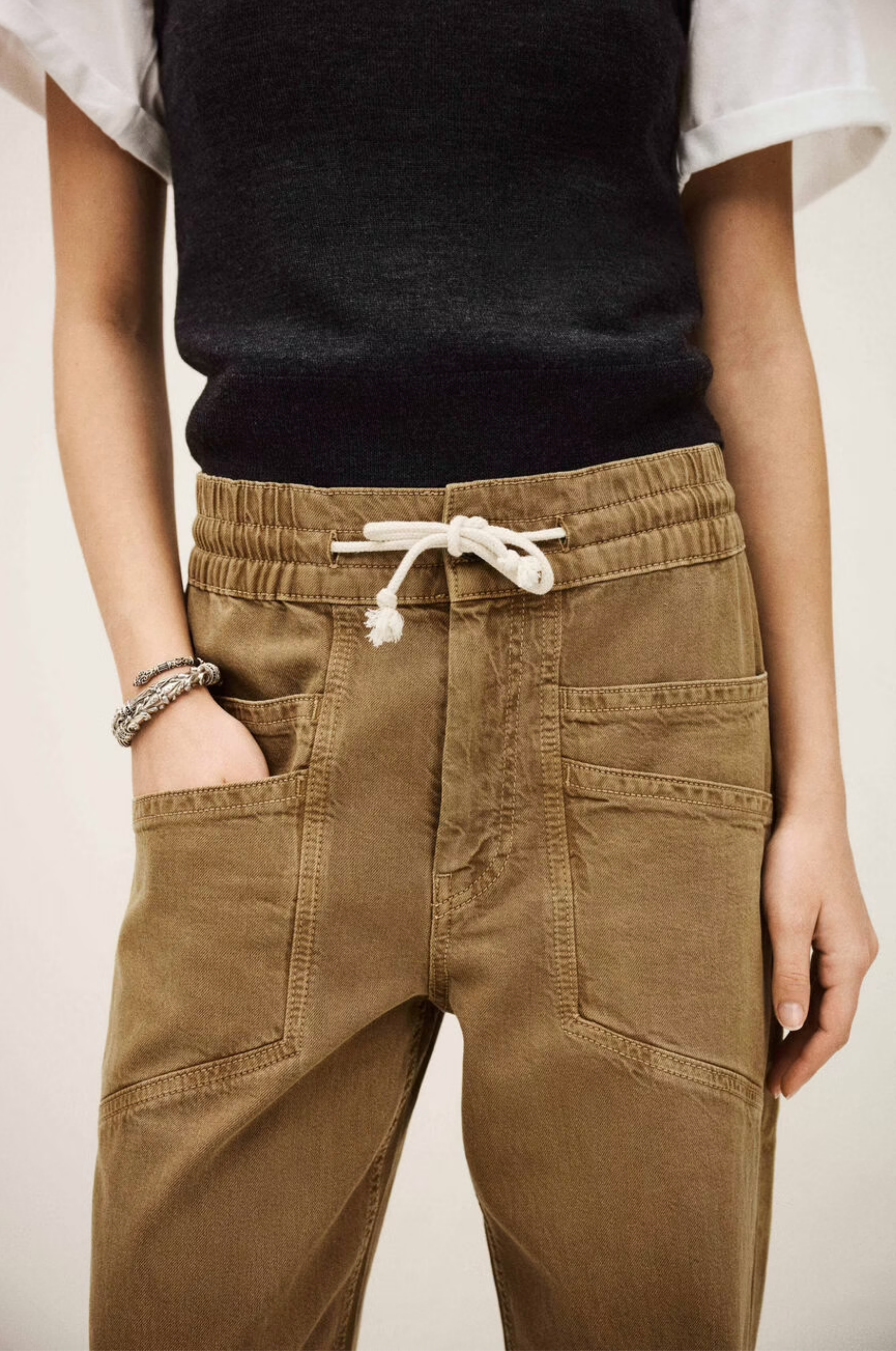 Wearing BA&SH Pant Fima—high waist brown drawstring pants with large front pockets—paired with a black sleeveless top over a white shirt and a silver bracelet, the person stands with one hand in their pocket.