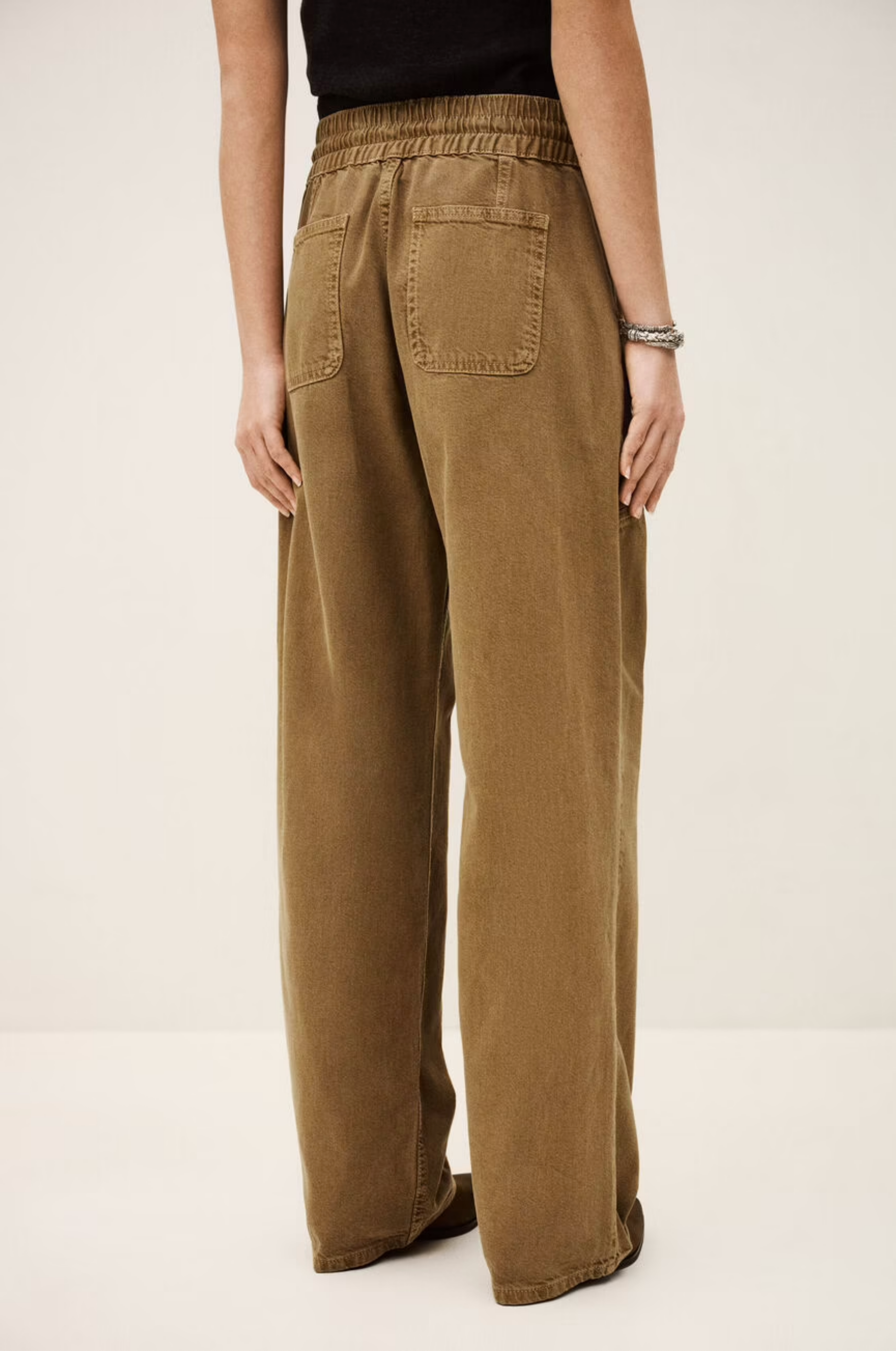 A person models the BA&SH Pant Fima—loose-fitting, high-waisted brown cotton linen pants with an elasticated waist and back pockets—against a neutral background, showing only the lower half of the body.