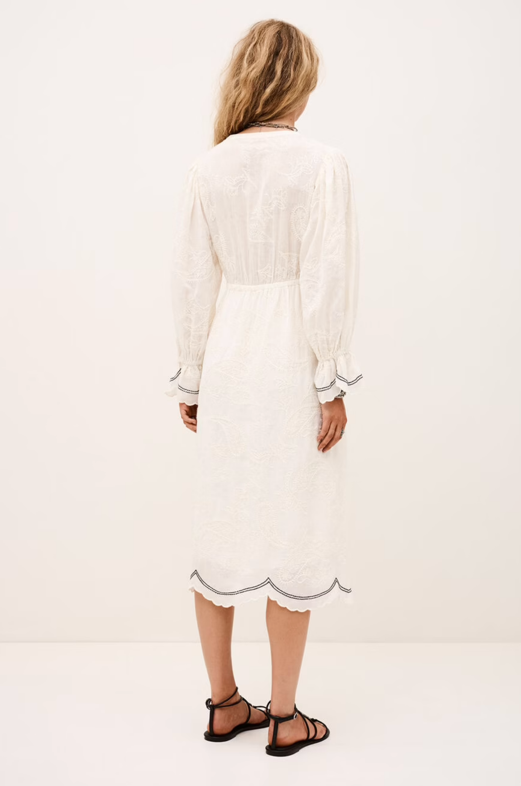 A woman with wavy blonde hair faces away, wearing the BA&SH Dress Prisco—a white, long-sleeved, knee-length lace dress with scalloped edges—paired with black sandals against a plain light background.