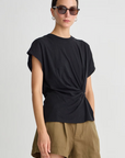 A woman wearing sunglasses, the Apiece Apart Volta Twist Top, and olive green shorts stands against a plain light gray background.
