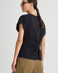 A woman with glasses and a braid wears the Apiece Apart Volta Twist Top, which features a gathered twist detail at the back, paired with olive green shorts against a plain light background.