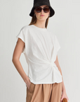 A woman in the Apiece Apart Volta Twist Top—a white organic cotton tee with a knot detail—black bucket hat, dark sunglasses, and loose tan pants stands against a plain light background, holding a brown item in her left hand.