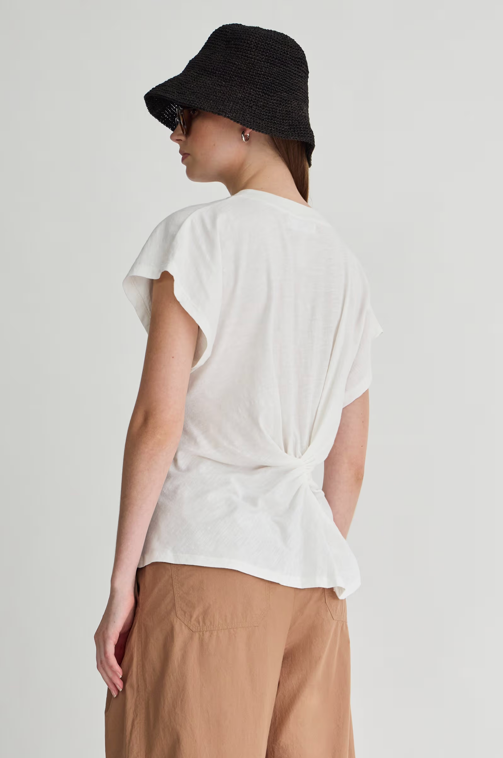 A person in the Apiece Apart Volta Twist Top, black bucket hat, sunglasses, and light brown shorts faces away from the camera against a plain background.