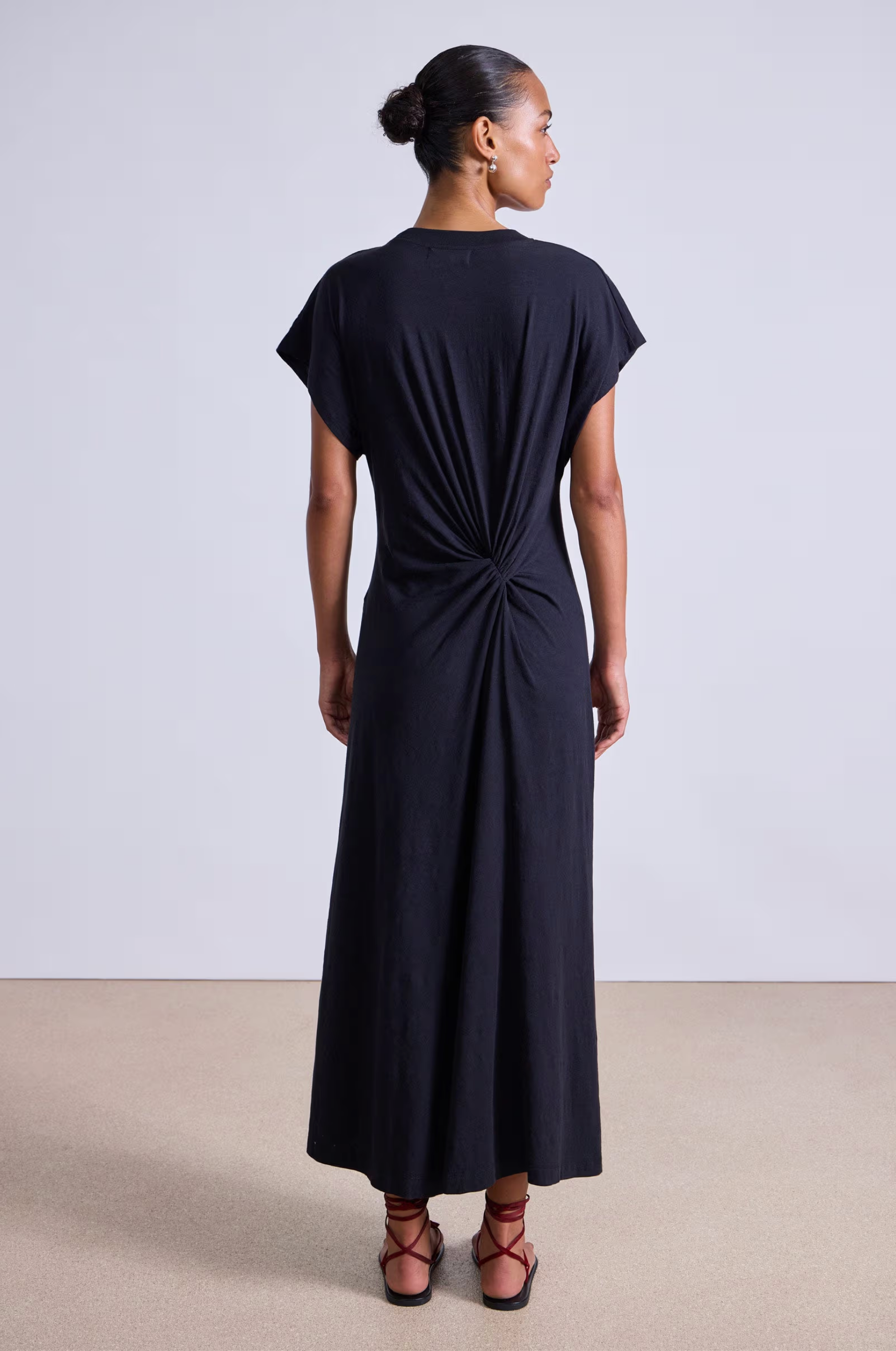 A woman stands facing away in the Apiece Apart Volta Twist Dress—a black organic cotton, ankle-length style with short sleeves and a gathered twist detail at the lower back. Her hair is in a bun, and she wears red strappy sandals against a plain background.