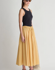 A woman wearing a black sleeveless top, sunglasses, black sandals, a black headband, and the Apiece Apart Hanne Tie Maxi Skirt in yellow stands against a plain background holding a netted bag.